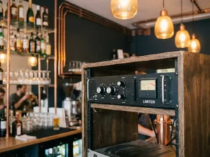 Zwarte audio limiter in rackmount achter een cafébalie, met warme amber hanglampen en glazen flessen op de achtergrond.