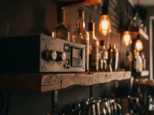 SPL-limiter op een verweerde houten barplank in een café, met rode en groene LED-indicatoren en wazige flessen op de achtergrond.