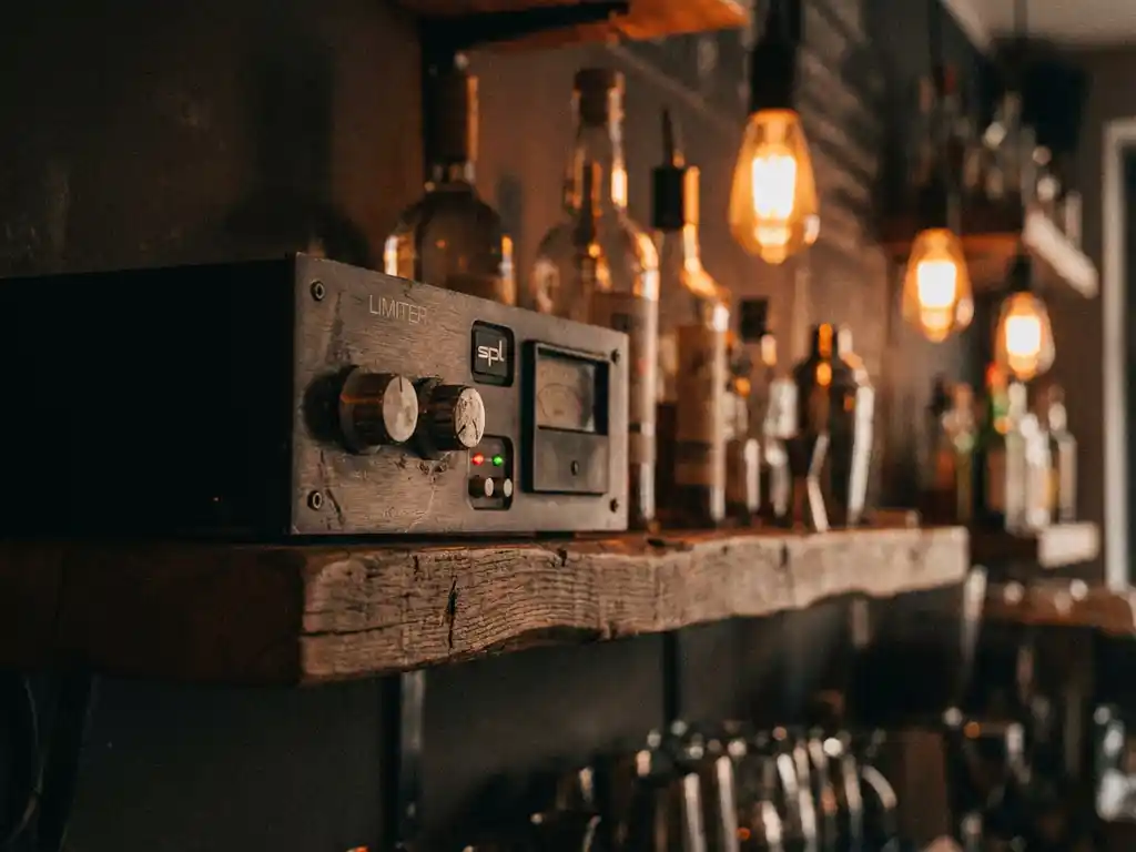 SPL-limiter op een verweerde houten barplank in een café, met rode en groene LED-indicatoren en wazige flessen op de achtergrond.