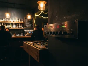 Zwarte audio limiter gemonteerd naast een DJ-booth in een café, met gloeiende LED-indicatoren en warme amber barverlichting op de achtergrond.