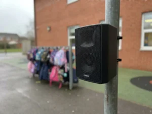 Zwarte LYNX passieve buitenluidspreker op stalen paal bij Nederlands schoolplein, rode bakstenen muur en wazige kinderrugzakken op achtergrond.