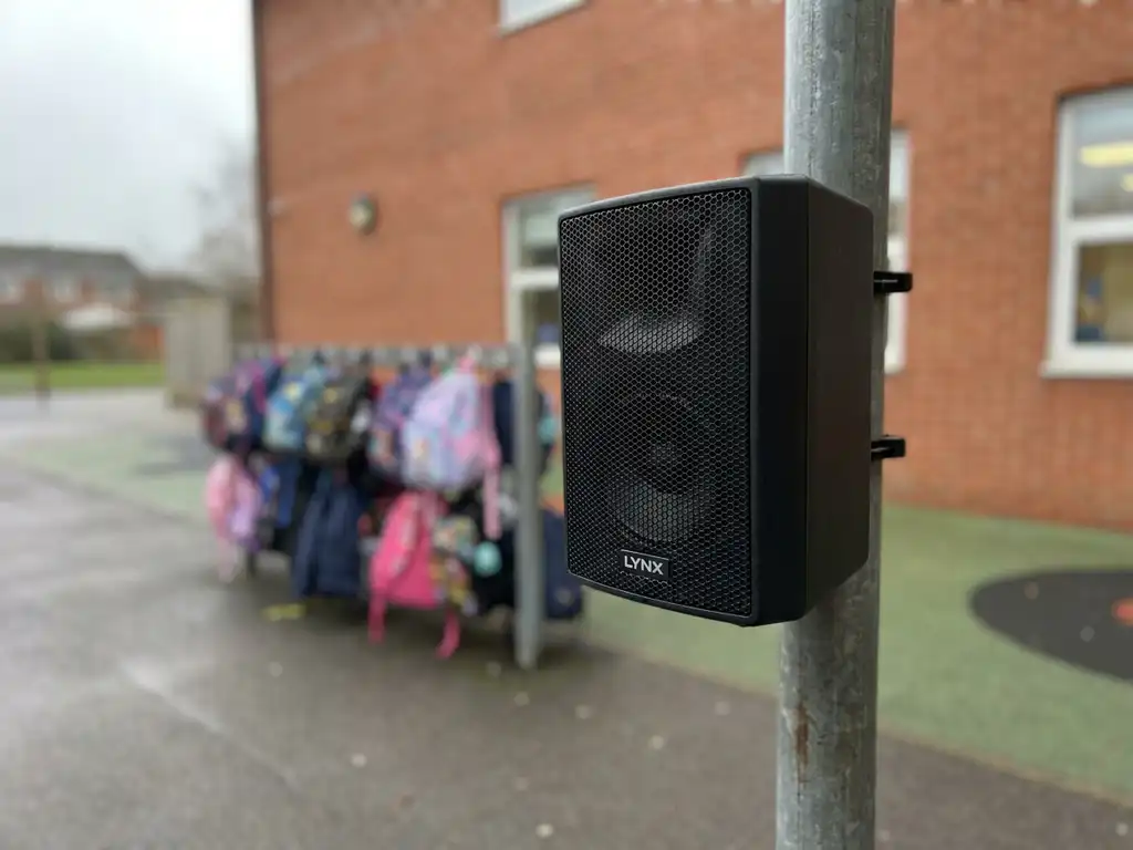 Zwarte LYNX passieve buitenluidspreker op stalen paal bij Nederlands schoolplein, rode bakstenen muur en wazige kinderrugzakken op achtergrond.