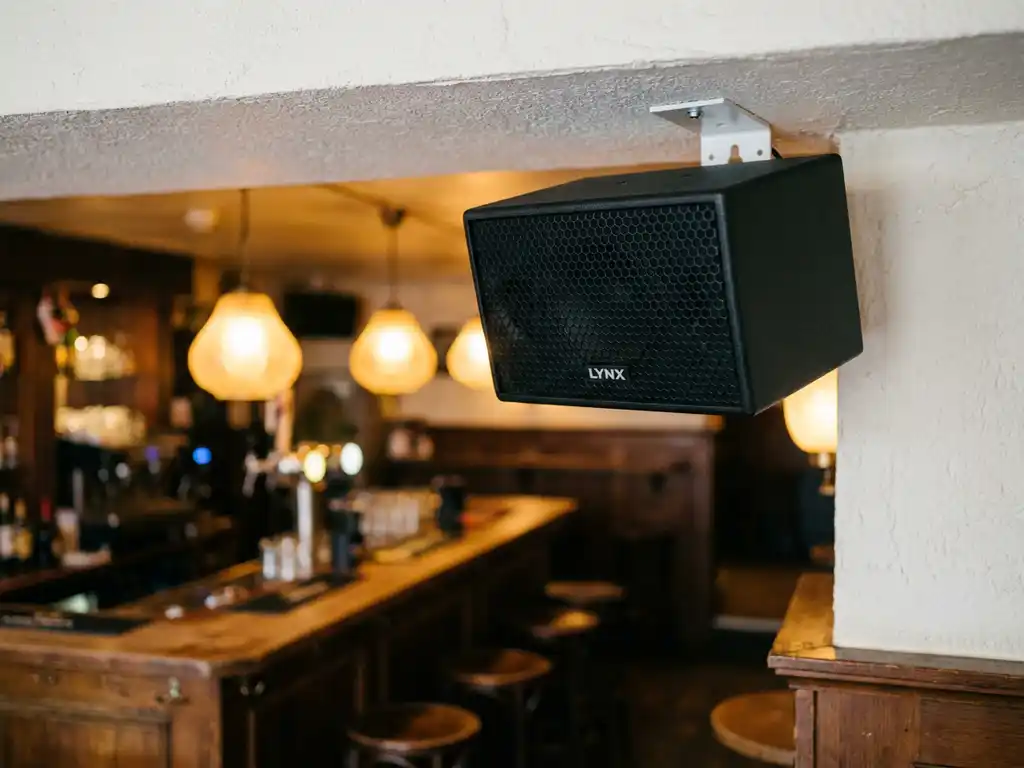 LYNX passieve speaker op witte plafondbeugel in gezellig Nederlands café met houten bar en warme amberkleurige hanglampen.