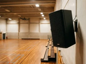 Compacte zwarte LYNX luidspreker gemonteerd aan de muur van een Nederlandse sporthal met houten vloer en opgevouwen tribunes.