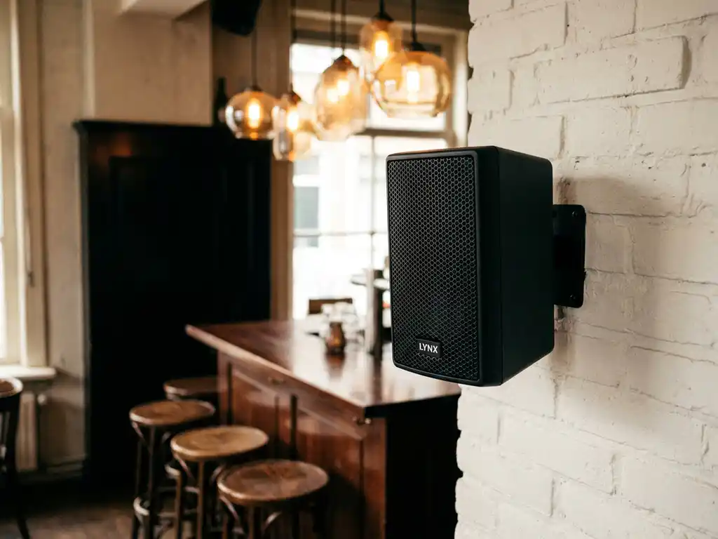 Compacte zwarte LYNX passieve speaker op witte bakstenen muur in Nederlands café, warme sfeerverlichting en houten barkrukken op achtergrond.