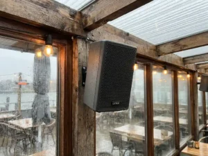 Compacte zwarte LYNX passieve speaker op verweerde houten balk in Nederlands terrasbar, regendruppels op glazen panelen op de achtergrond.