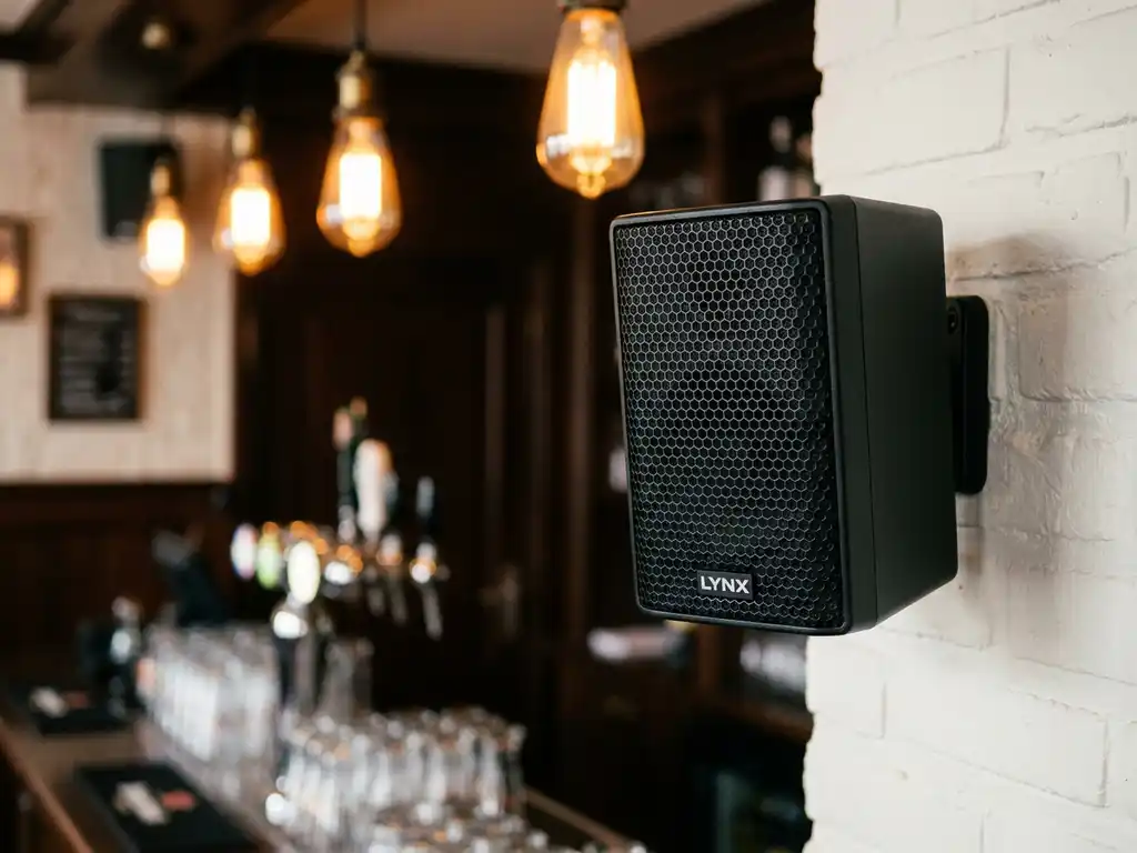 Compacte zwarte LYNX speaker met hexagonaal metalen rooster op witte bakstenen muur in een gezellig bruin café.