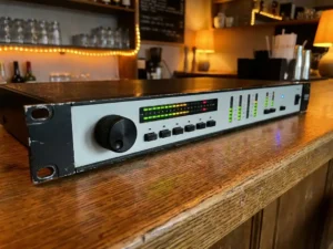 Zwarte 1U rack-mount audio limiter met spectrumdisplay en LED-meters op een verweerde eiken bartafel in een sfeervol café.