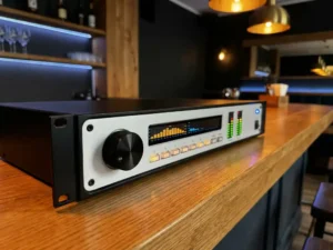 1U rack-mount audio limiter op een eiken bartafel in een restaurant, met draaiknop, spectrumanalyzer en blauwe LED-indicator.