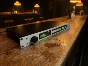 Zwarte 1U rack-mount audio limiter op een versleten eiken bartafel, met draaiknop, spectrumanalyzer en blauwe LED, warm pub-licht.