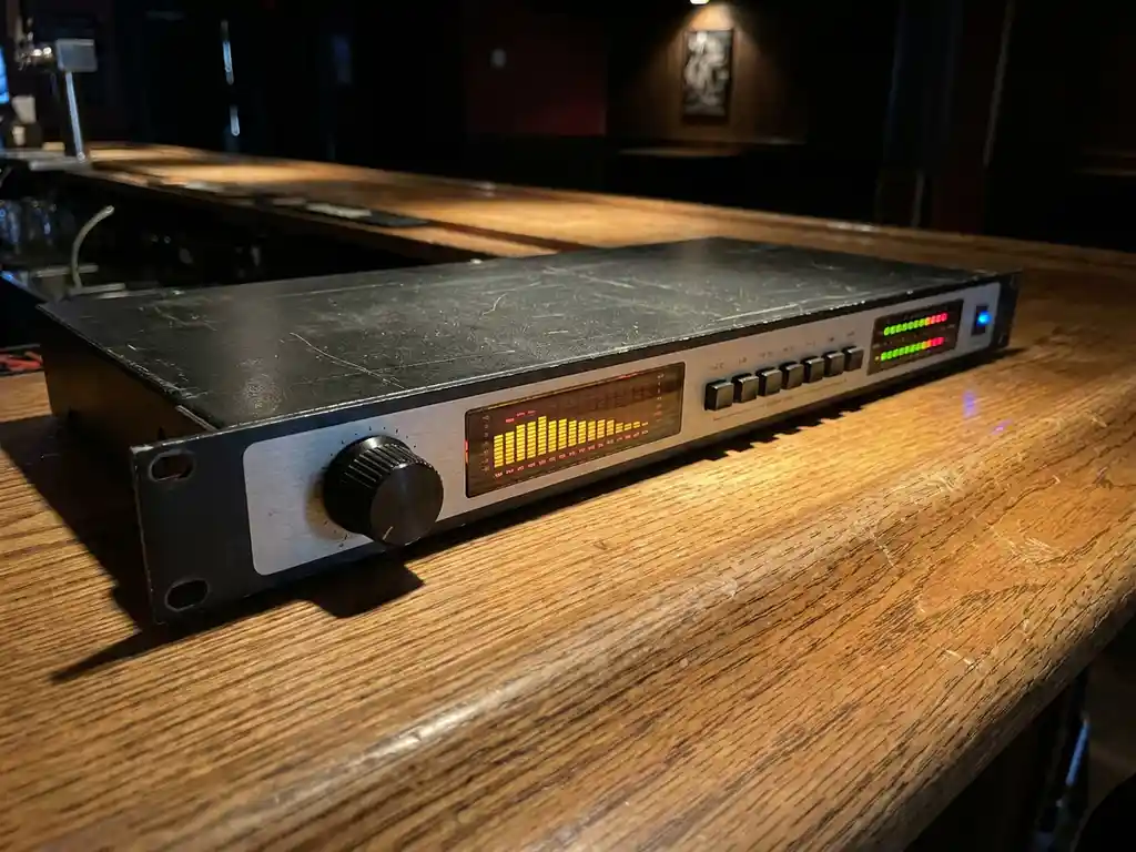 Zwarte 1U rack-mount audiolimiter met roterende knop, spectrumanalysator en LED-meters op een versleten eiken bardek in een donkere horecagelegenheid.
