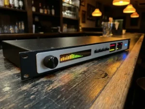 1U rack-mount audiolimiter met spectrumdisplay en LED-meters op een verweerde eiken bartafel, verlicht door warm amberkleurig publicht.