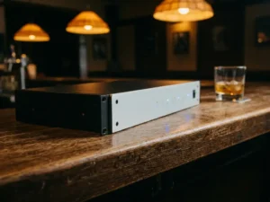 Zwarte 1U rack-unit met lichtgrijs front panel op een eiken bartafel, blauw gloeiende LED-indicatoren, whiskeyglas op de achtergrond.